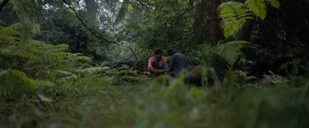 Movie still from “In the Earth” (2021), directed by Ben Wheatley – Two people sitting in the woods near some trees; Wide shot, Over the shoulder angle