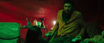 Movie still from “In the Earth” (2021), directed by Ben Wheatley – A man sitting in front of a woman in a green room; Medium shot, High angle