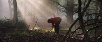 Movie still from “In the Earth” (2021), directed by Ben Wheatley – A man in a red jacket is digging in the woods; Wide shot, Low angle
