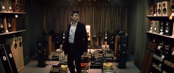 Movie still from “Infernal Affairs” (2002), directed by Alan Mak – A man in a suit standing in front of a room full of audio equipment; Medium shot, Low angle