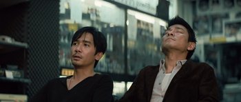 Movie still from “Infernal Affairs” (2002), directed by Alan Mak – Two asian men sitting next to each other in front of a building; Medium shot, Low angle