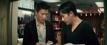 Movie still from “Infernal Affairs” (2002), directed by Alan Mak – Two asian men looking at a book in a store; Medium shot, Low angle