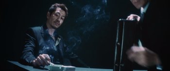 Movie still from “Infernal Affairs” (2002), directed by Alan Mak – A man sitting at a table in front of a laptop computer with smoke coming out of his mouth; Medium shot, Low angle