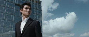 Movie still from “Infernal Affairs” (2002), directed by Alan Mak – A man wearing a suit and tie standing in front of a building; Medium shot, Low angle