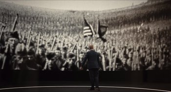 Movie still from “Inferno” (2016), directed by Ron Howard – A man standing in front of an image of a crowd of people holding american flags; Wide shot, High angle