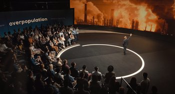 Movie still from “Inferno” (2016), directed by Ron Howard – An audience is watching a man on a stage in front of an image of an oil refinery; Extreme Wide shot, High angle