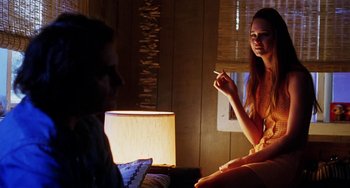 Movie still from “Inherent Vice” (2014), directed by Paul Thomas Anderson – A woman smoking a cigarette in front of a man in front of a lamp; Medium shot, Over the shoulder angle