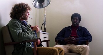 Movie still from “Inherent Vice” (2014), directed by Paul Thomas Anderson – Two people sitting in a room with a lamp on the wall behind them; Medium shot, Over the shoulder angle