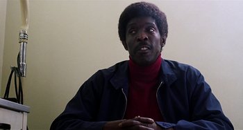 Movie still from “Inherent Vice” (2014), directed by Paul Thomas Anderson – A black man with a red shirt is sitting down; Close Up shot, Over the shoulder angle