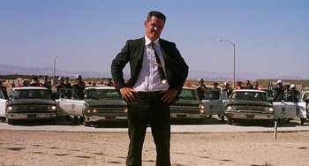 Movie still from “Inherent Vice” (2014), directed by Paul Thomas Anderson – A man in a suit and tie standing in front of cars; Wide shot, Low angle