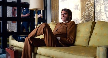 Movie still from “Inherent Vice” (2014), directed by Paul Thomas Anderson – A man sitting on top of a couch in a room; Medium shot, Low angle