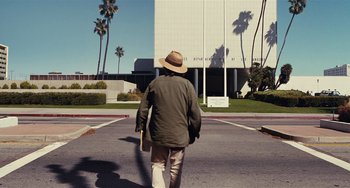 Movie still from “Inherent Vice” (2014), directed by Paul Thomas Anderson – A man with a hat is walking down the street with a skateboard; Wide shot, Low angle