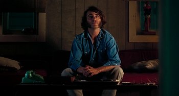 Movie still from “Inherent Vice” (2014), directed by Paul Thomas Anderson – A man sitting on a red couch in a room; Medium shot, Low angle