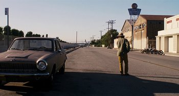 Movie still from “Inherent Vice” (2014), directed by Paul Thomas Anderson – A man standing on the side of a road next to a car; Wide shot, Over the shoulder angle