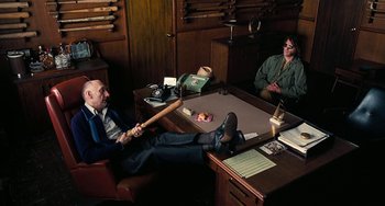Movie still from “Inherent Vice” (2014), directed by Paul Thomas Anderson – Two men sitting at a table with a bat; Wide shot, High angle