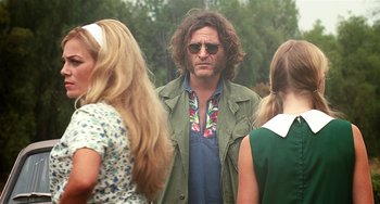 Movie still from “Inherent Vice” (2014), directed by Paul Thomas Anderson – A man with long hair wearing sunglasses standing next to two other women; Medium shot, Over the shoulder angle
