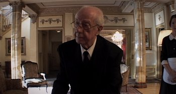 Movie still from “Inland Empire” (2006), directed by David Lynch – An older man in a suit and tie in a room; Medium shot, Low angle