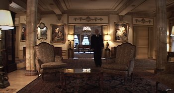 Movie still from “Inland Empire” (2006), directed by David Lynch – A man in a suit and tie standing in front of two chairs; Wide shot, Low angle