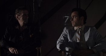 Movie still from “Inland Empire” (2006), directed by David Lynch – A man in a suit and tie standing next to another man in a suit and tie; Medium shot, Over the shoulder angle