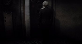 Movie still from “Inland Empire” (2006), directed by David Lynch – A woman standing in front of an open door in a dark room; Wide shot, High angle