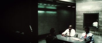 Movie still from “Inside Man” (2006), directed by Spike Lee – Two men are sitting at a table in a dark room; Wide shot, Over the shoulder angle