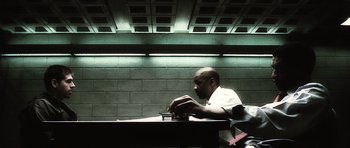 Movie still from “Inside Man” (2006), directed by Spike Lee – A man sitting at a table in front of a brick wall; Medium shot, Over the shoulder angle