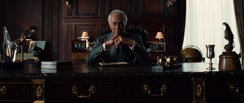 Movie still from “Inside Man” (2006), directed by Spike Lee – An older man sitting at a desk in front of a book; Medium shot, Low angle