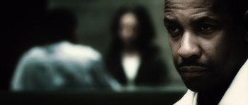 Movie still from “Inside Man” (2006), directed by Spike Lee – A man is looking at the camera while a woman is sitting in the background; Extreme Close Up shot, Over the shoulder angle