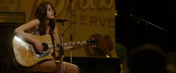 Movie still from “Into the Wild” (2007), directed by Sean Penn – A woman sitting on a stage with a microphone and a guitar; Medium shot, Over the shoulder angle