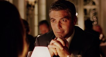 Movie still from “Intolerable Cruelty” (2003), directed by Joel Coen – A man sitting at a table with his hands clasped together; Close Up shot, Over the shoulder angle