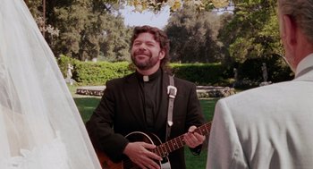 Movie still from “Intolerable Cruelty” (2003), directed by Joel Coen – A man in a priest's outfit holding a guitar; Medium shot, Over the shoulder angle