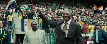 Movie still from “Invictus” (2009), directed by Clint Eastwood – An older man in a suit and tie holding his hand up in front of a crowd; Medium shot, Low angle