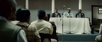 Movie still from “Invictus” (2009), directed by Clint Eastwood – A group of people sitting at a table with microphones and microphones; Medium shot, Over the shoulder angle