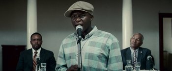 Movie still from “Invictus” (2009), directed by Clint Eastwood – A man in a plaid shirt is speaking into a microphone; Close Up shot, Low angle