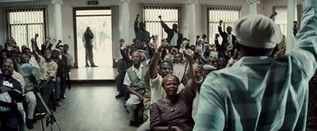 Movie still from “Invictus” (2009), directed by Clint Eastwood – A group of people sitting in a room with their hands raised; Wide shot, High angle