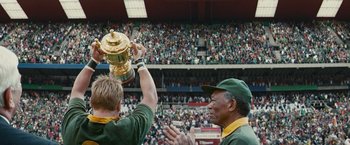 Movie still from “Invictus” (2009), directed by Clint Eastwood – A man holding up a trophy in front of a crowd; Medium shot, Low angle