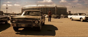 Movie still from “Invincible” (2006), directed by Ericson Core – A man is standing in front of an old car in a parking lot; Wide shot, Low angle