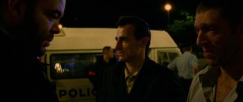 Movie still from “Irreversible” (2002), directed by Gaspar Noé – A man wearing a suit and tie standing in front of a police car; Close Up shot, Over the shoulder angle