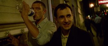 Movie still from “Irreversible” (2002), directed by Gaspar Noé – Two men standing next to each other and one of them is drinking a beer; Medium shot, Low angle
