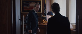 Movie still from “Jason Bourne” (2016), directed by Paul Greengrass – A man and a woman in a room with a flag on the wall behind them; Medium shot, Over the shoulder angle