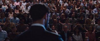 Movie still from “Jason Bourne” (2016), directed by Paul Greengrass – A crowd of people sitting and standing in front of a speaker; Wide shot, Over the shoulder angle