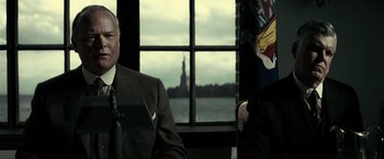 Movie still from “J. Edgar” (2011), directed by Clint Eastwood – A man in a suit sitting next to another man in front of a statue of liberty; Medium shot, Over the shoulder angle