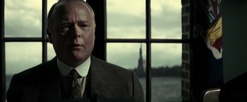 Movie still from “J. Edgar” (2011), directed by Clint Eastwood – An older man wearing a suit and tie in front of the statue of liberty in new york city; Close Up shot, Over the shoulder angle