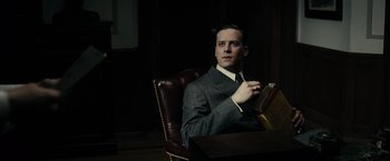 Movie still from “J. Edgar” (2011), directed by Clint Eastwood – A man sitting in a chair holding an open book; Medium shot, Low angle