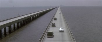 Movie still from “JFK” (1991), directed by Oliver Stone – Cars are driving on a bridge over a body of water; Extreme Wide shot, High angle
