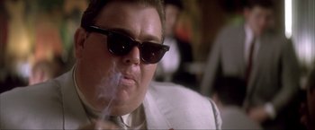 Movie still from “JFK” (1991), directed by Oliver Stone – A man with sunglasses smoking a cigarette in front of a group of people; Close Up shot, Low angle