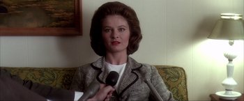 Movie still from “JFK” (1991), directed by Oliver Stone – A woman is holding a microphone while sitting on a couch; Close Up shot, Over the shoulder angle