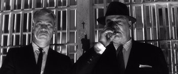 Movie still from “JFK” (1991), directed by Oliver Stone – A man smoking a cigarette next to another man in a suit and tie; Close Up shot, Low angle