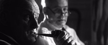 Movie still from “JFK” (1991), directed by Oliver Stone – An older man smoking a pipe while another man watches; Close Up shot, Low angle