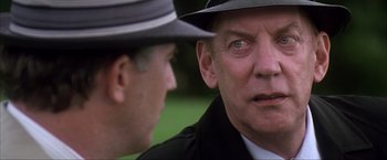 Movie still from “JFK” (1991), directed by Oliver Stone – A man in a suit and a hat talking to another man in a hat; Close Up shot, Over the shoulder angle